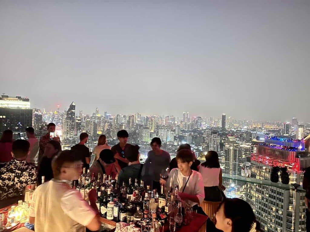 Bangkok: Barkeeper an der Tichuca Rooftop Bar mit Gästen und dem nächtlichen Bangkok im Hintergrund